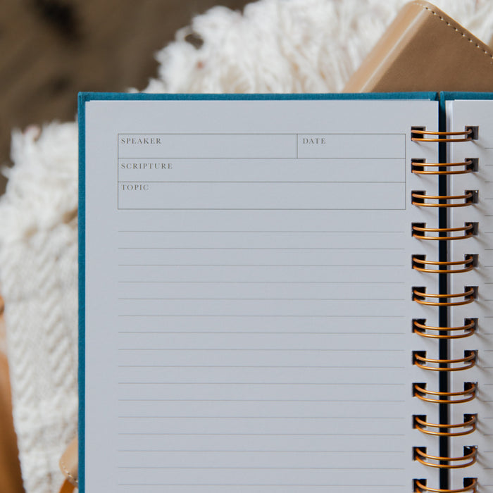 An open spiral-bound planner or notebook with lined pages, featuring fields for speaker, date, scripture, and topic. The notebook has a blue cover edge and gold-toned spiral binding, photographed on a soft white textured blanket with a tan leather-like object in the background.