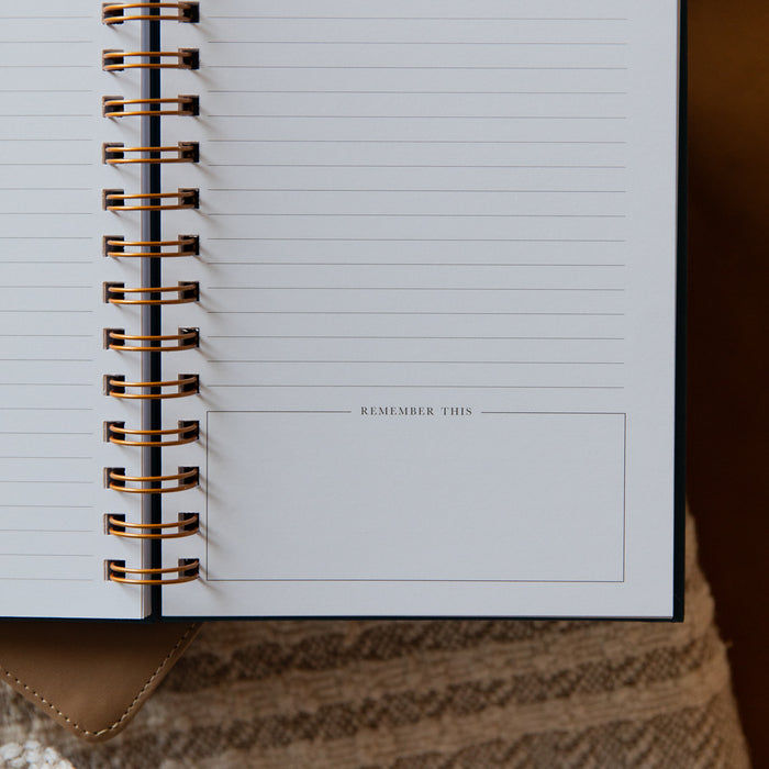 An open spiral-bound notebook with lined pages and a blank note section labeled 'REMEMBER THIS,' photographed close up with a warm neutral background.