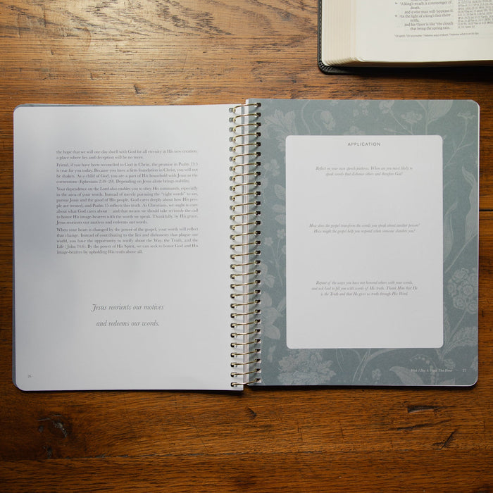 An open spiral-bound booklet set lies on a wooden tabletop, with one page showing printed text and the other featuring a large blank white card labeled 'APPLICATION.' A second open book is partially visible in the top right corner.
