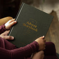 A person holding a dark green hardcover book titled 'The Advent Storybook' with gold lettering, shown in a cozy indoor setting near a Christmas tree with warm lights.