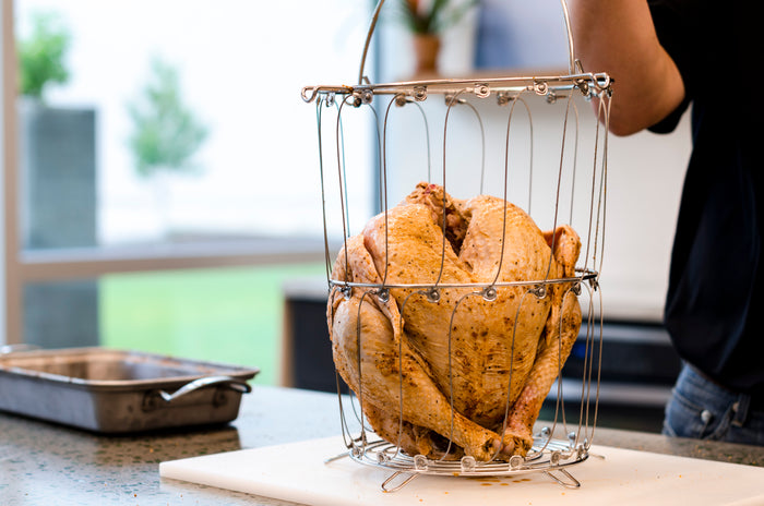 Silver wire roasting basket holding a whole seasoned chicken on a white cutting board.