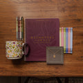 A styled flat-lay on a wooden table featuring a maroon spiral-bound journal with gold lettering that reads 'Daughters of Grace' and '150th Anniversary Edition.' Surrounding it are a floral ceramic mug, a set of metallic pens, pastel highlighters, and a small jewelry card holding a gold pendant necklace.