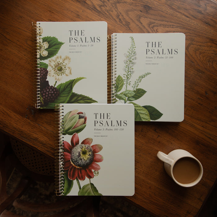 Three spiral-bound books titled "THE PSALMS" arranged on a wooden table, each with different botanical illustrations on the cover, plus a white mug of coffee nearby.
