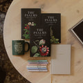 Three hardcover books titled 'The Psalms' arranged on a round light wood table, alongside a dark green ceramic mug with gold lettering, a stack of small pastel notebooks, and a cream-colored journal.
