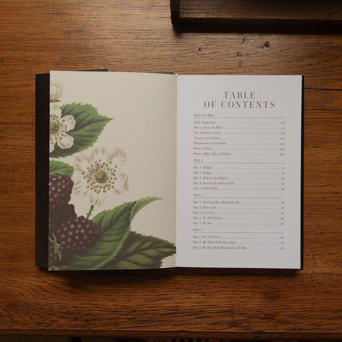 An open book laid flat on a wooden table, showing a table of contents page with a botanical illustration of white flowers and dark berries on the left page.