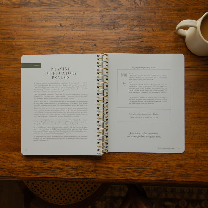 An open spiral-bound book titled 'Praying Imperative Psalms' lies on a wooden table, with a cream ceramic mug partially visible in the top right corner.