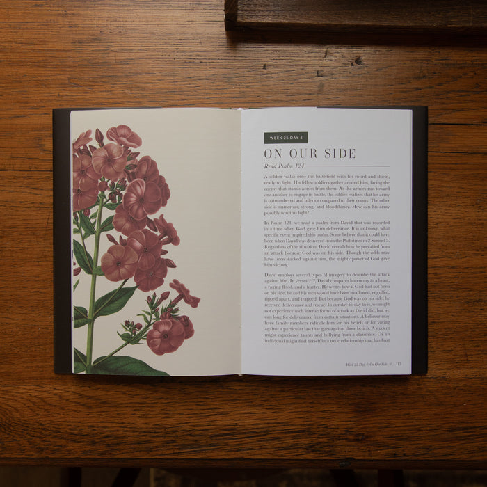 An open book lying flat on a wooden table, showing a botanical illustration of pink flowers on the left page and a text article titled 'ON OUR SIDE' on the right page.