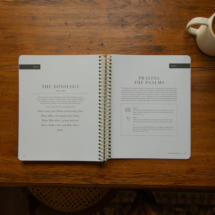 An open spiral-bound book laid flat on a wooden table, showing printed pages titled 'The Donology' and 'Praying the Psalms.' A small cream-colored ceramic cup is partially visible in the top-right corner.