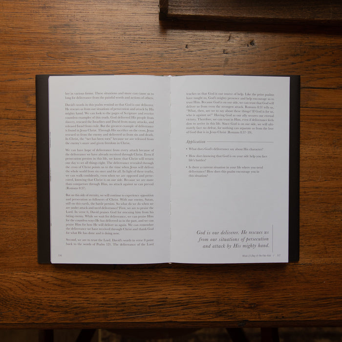 An open Bible book laid flat on a dark wooden table, showing two open pages with printed text and a small prayer at the bottom right of the right page.